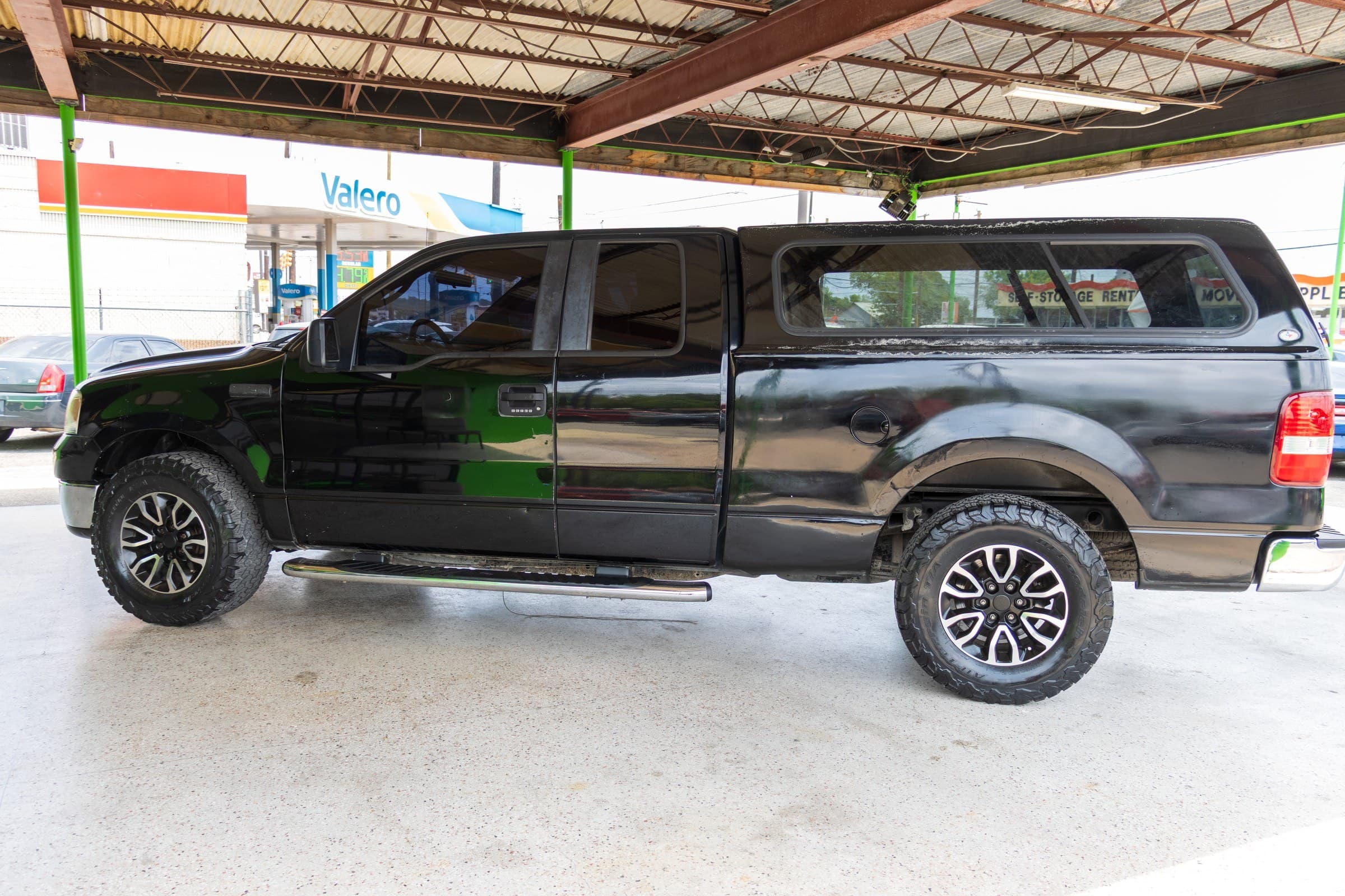 Full driver-side profile of the black F-150 with the camper shell fitted, parked under the lot canopy.