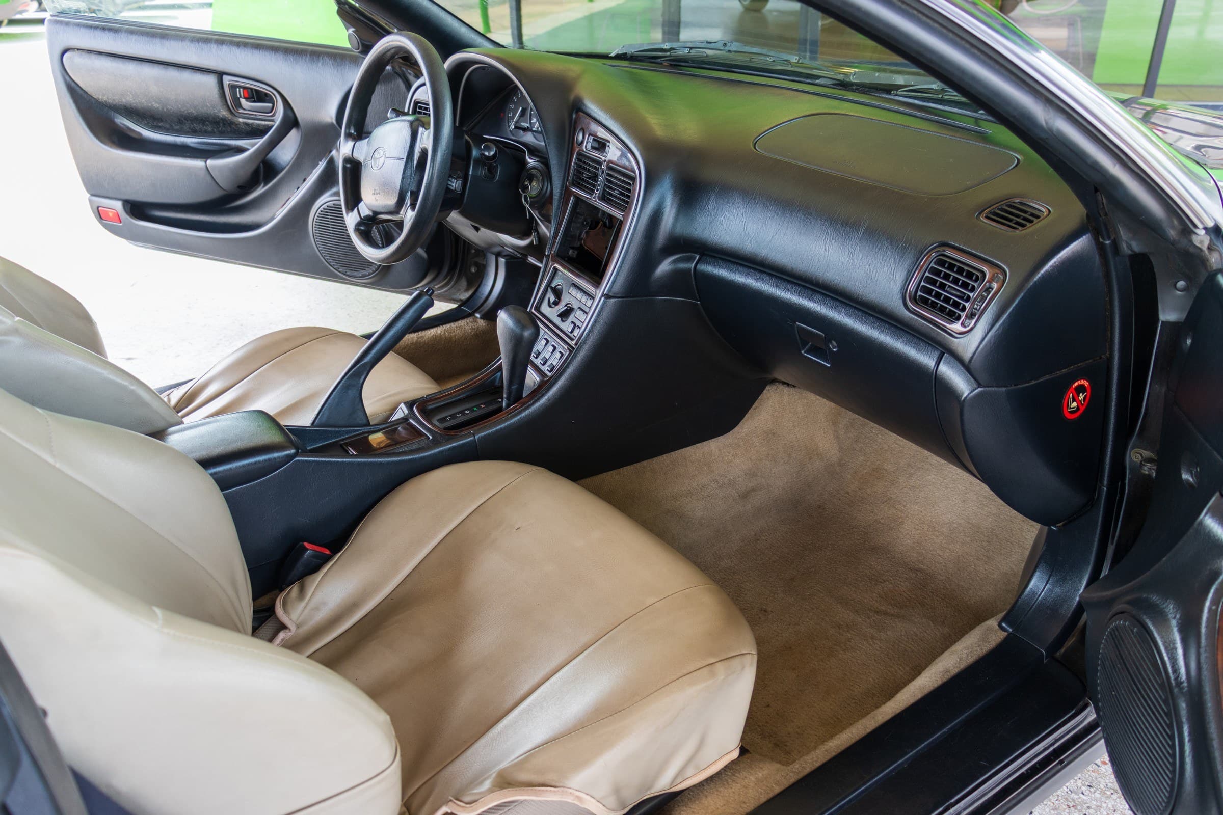 Celica interior seen through the open passenger door, showing the automatic shifter, wood-grain console and tan leather seats.