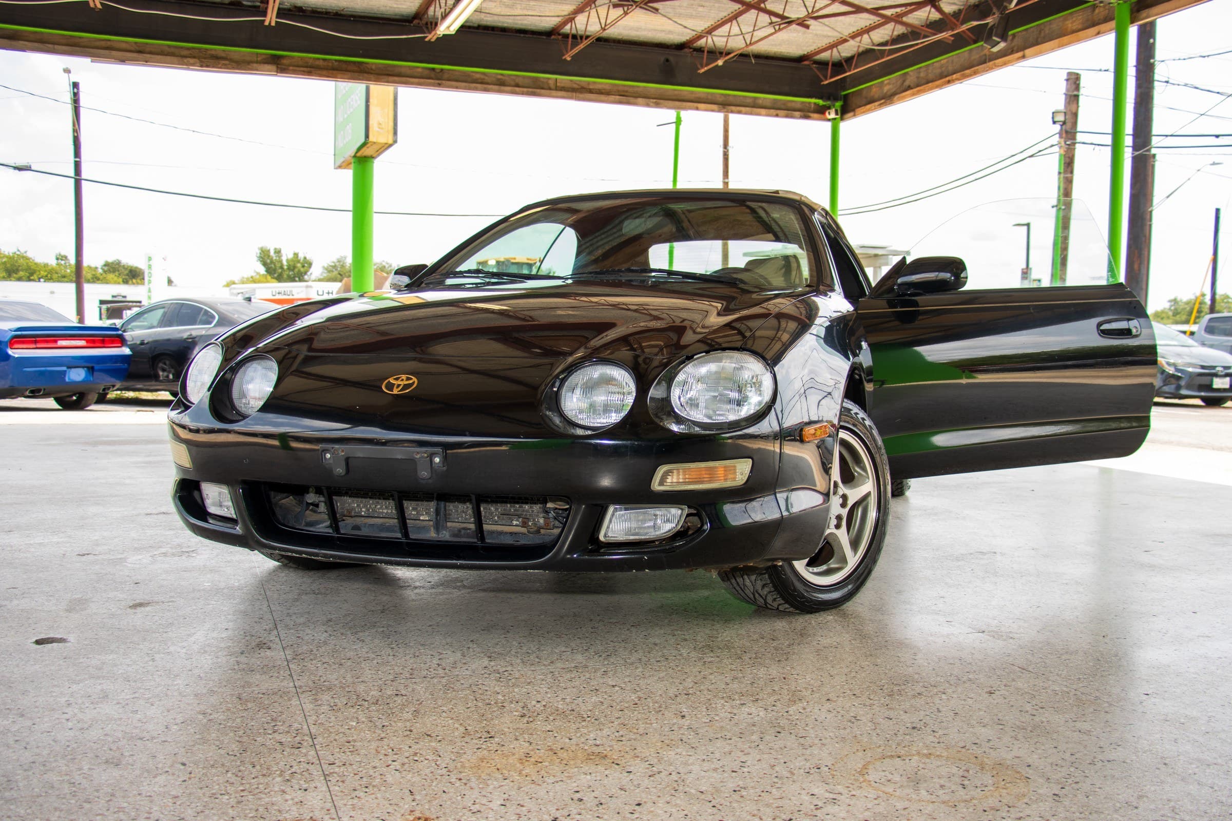 Low front view of the black Celica convertible with the passenger door open, showing its paired round headlights and gold Toyota badge.