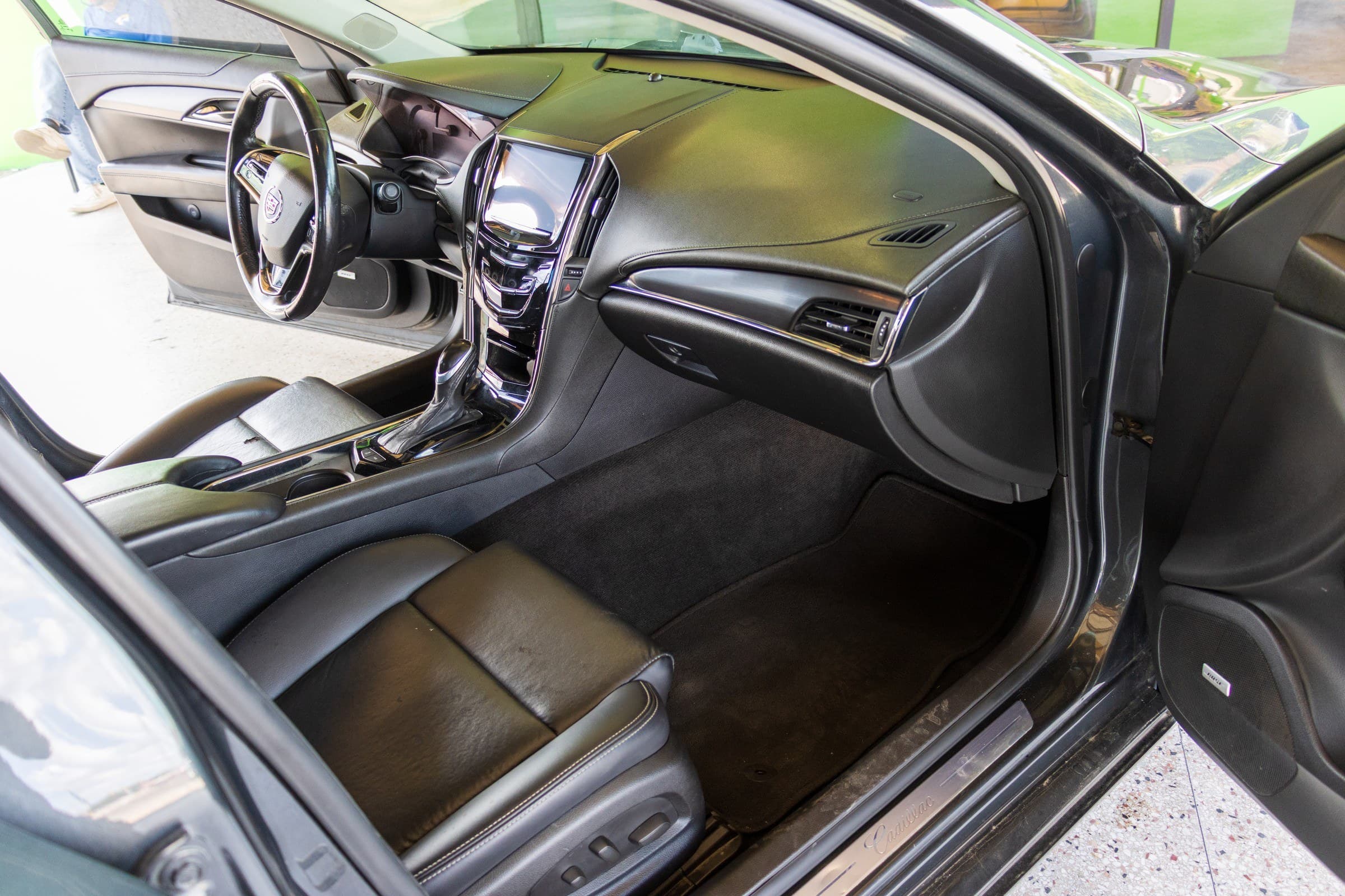 ATS cabin seen through the open passenger door, showing the CUE touchscreen, black leather seats and a Bose speaker in the door.